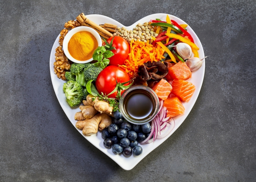 Heart healthy foods, good for lowering cholesterol, in a heart-shaped dish including acai, lentils, soy sauce, ginger, salmon, carrot, tomato, turmeric, cinnamon, walnuts, garlic, peppers, broccoli, basil, onion