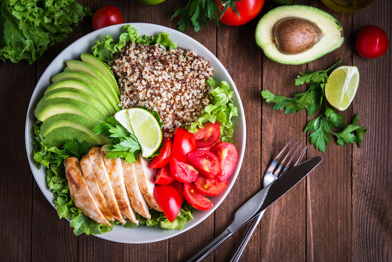 Bowl with leafy greens, quinoa, chicken, tomato, and lime wedges sit on top of a wooden table with silverware.