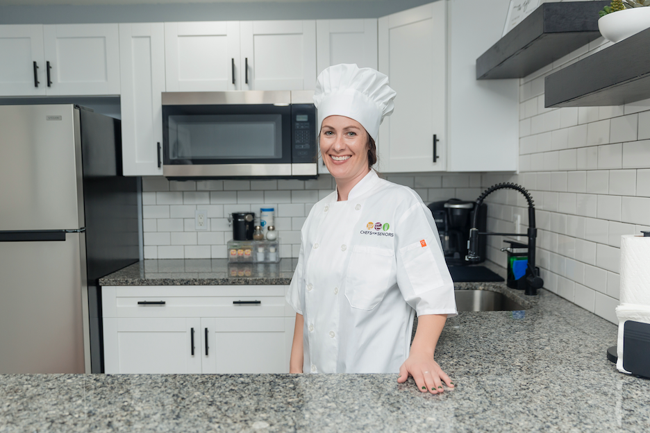 Personal Chef Jen wears a white chef coat with Chefs For Seniors logo and white chef hat, standing in kitchen