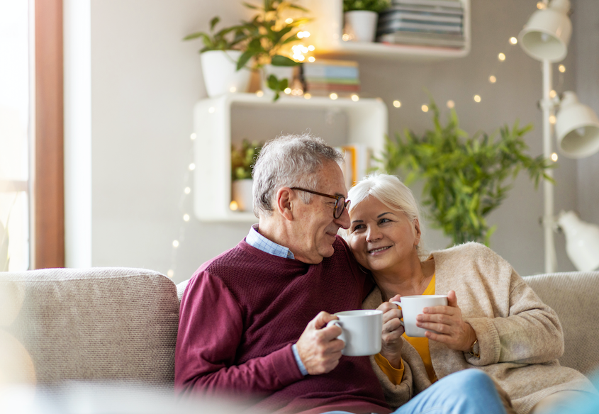 senior couple smiles at each other while cuddling on the couch holding white coffee mugs