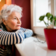 Senior woman with arms folded on window sill and chin resting on folded arms looks out the window, appears lonely