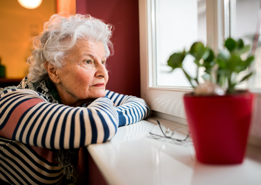 Senior woman with arms folded on window sill and chin resting on folded arms looks out the window, appears lonely
