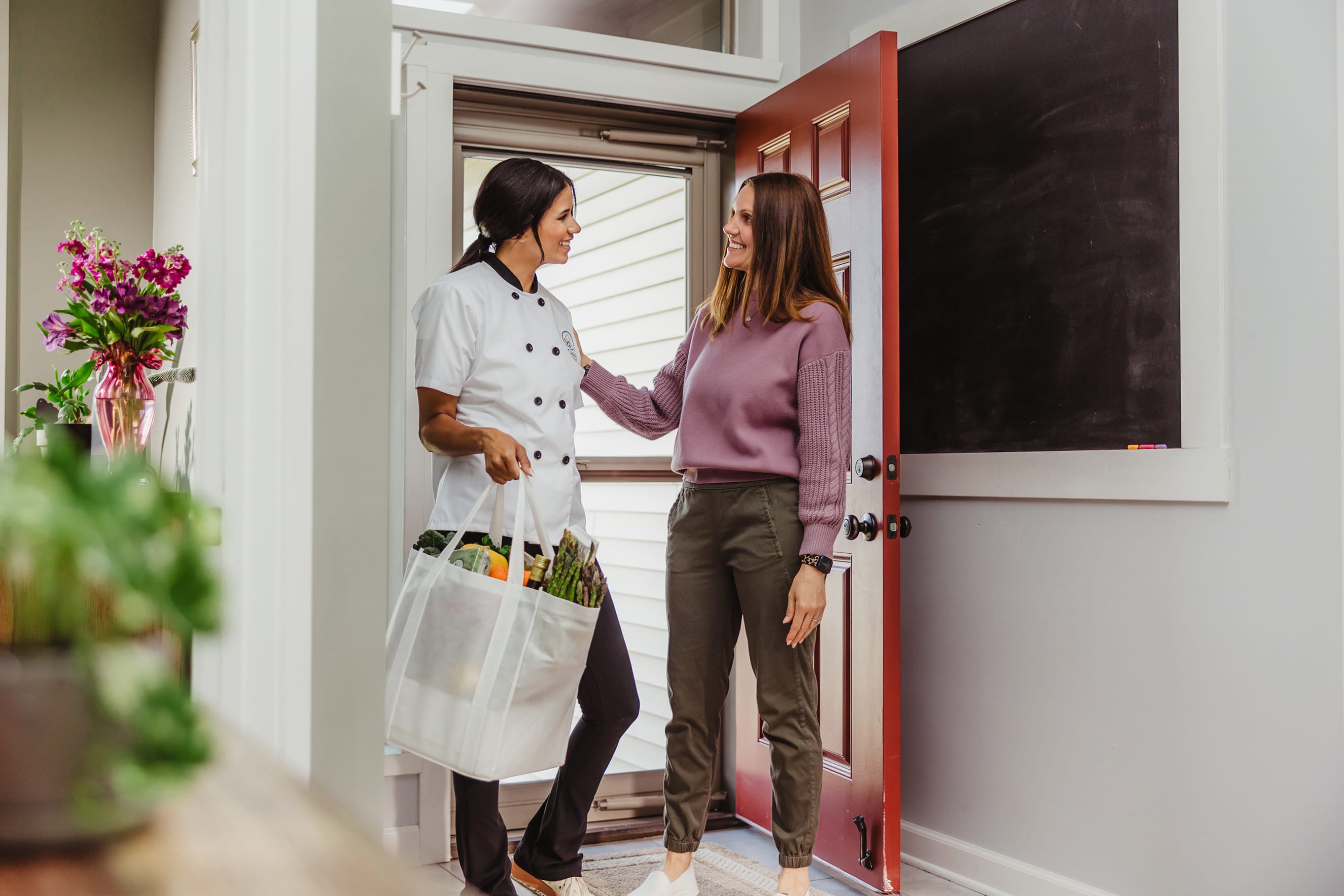 Chef carrying a bag of fresh groceries greeted by a woman at the front door