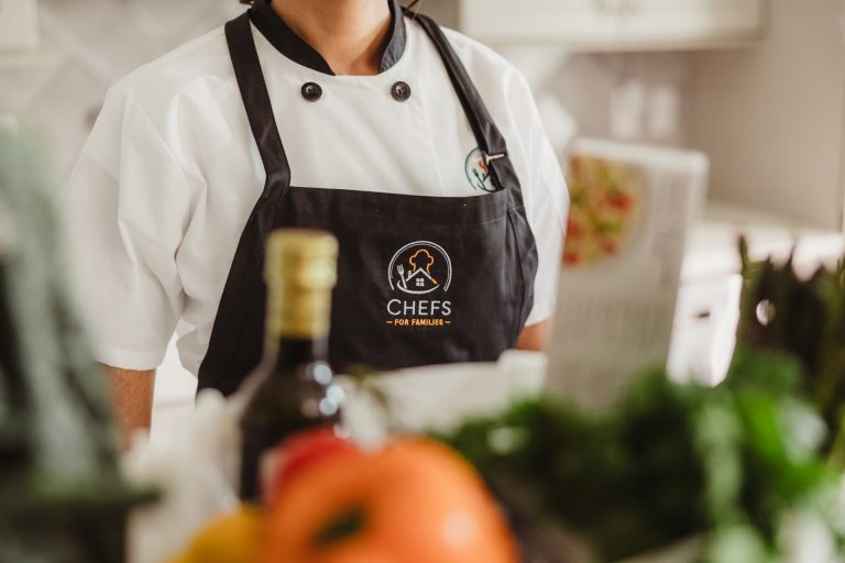 Chef in a Chefs for Families apron standing behind fresh produce and olive oil in a kitchen