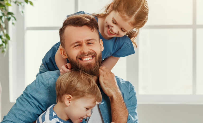 Bearded man laughing with a young boy and girl climbing on him at home